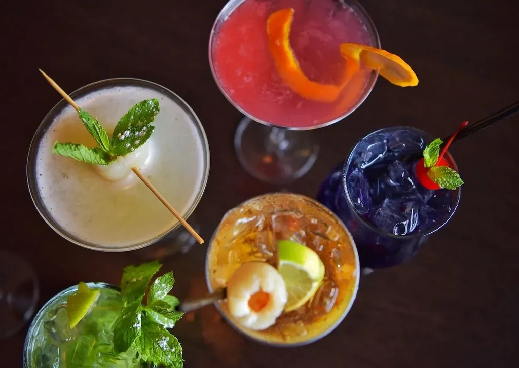 Overhead view of five distinct, colorful, garnished cocktails on dark surface.