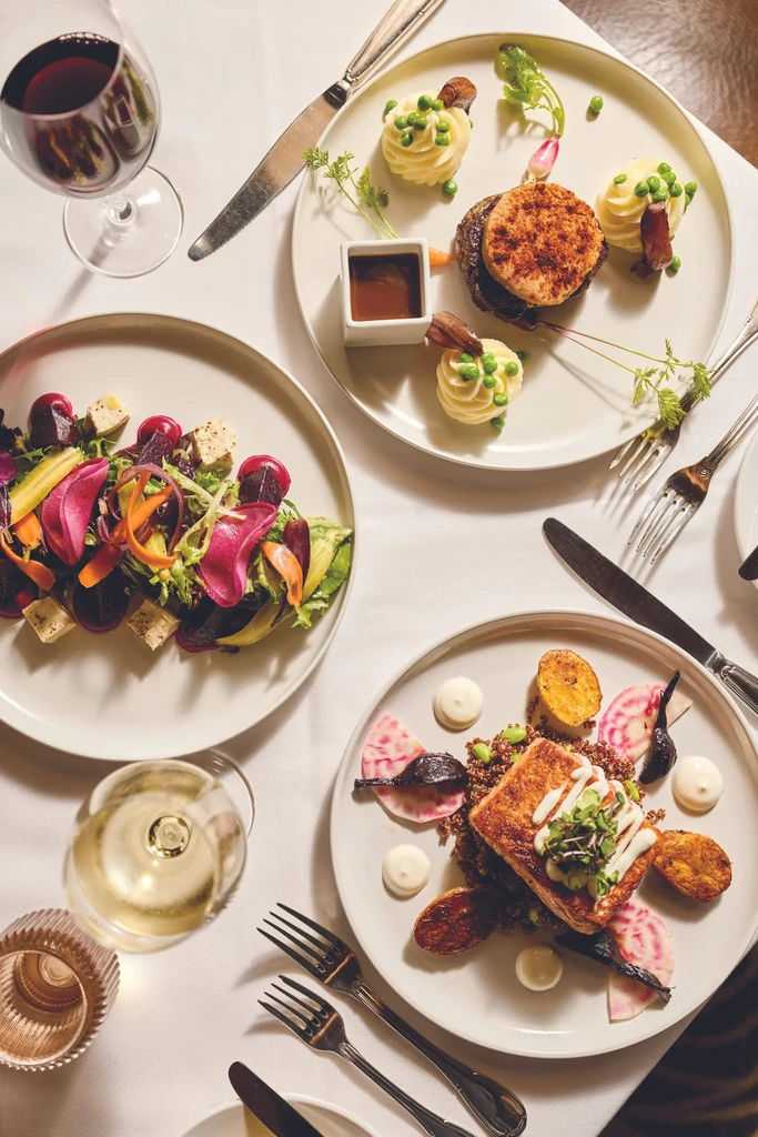 Three artistically plated dishes and wine on a white-clothed dining table.