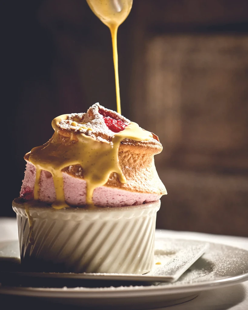 Raspberry soufflé in ramekin with powdered sugar and pouring custard sauce.