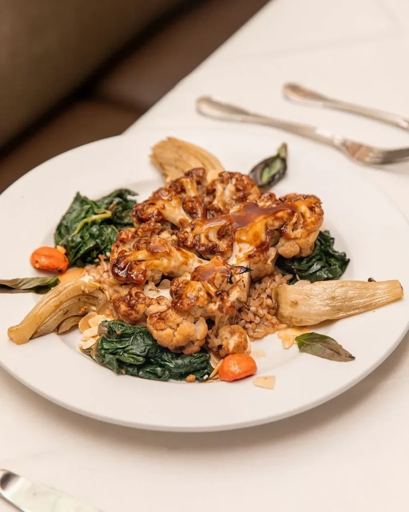 Roasted cauliflower, dark sauce, greens, grains, fennel, and almonds on a plate.