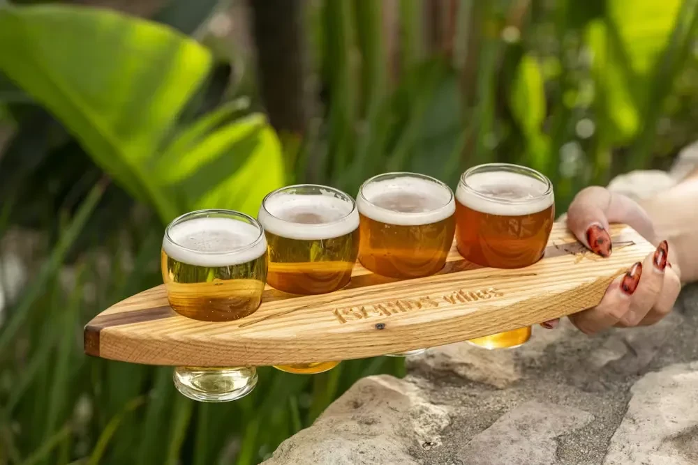 Person holds four-beer flight on wooden paddle outdoors.