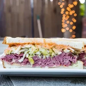 Toasted pastrami sandwich with coleslaw on a plate, blurred string lights.