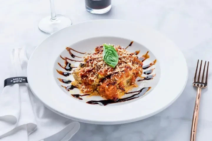 Layered pasta bake with cheese, basil, fork, and St. BAILY'S napkin.