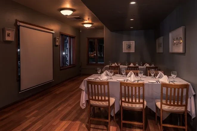 Dimly lit restaurant interior with set tables, wooden floors, projector screen.