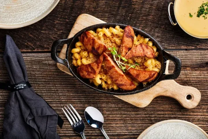 Overhead mac and cheese with grilled sausage in skillet on wooden board.