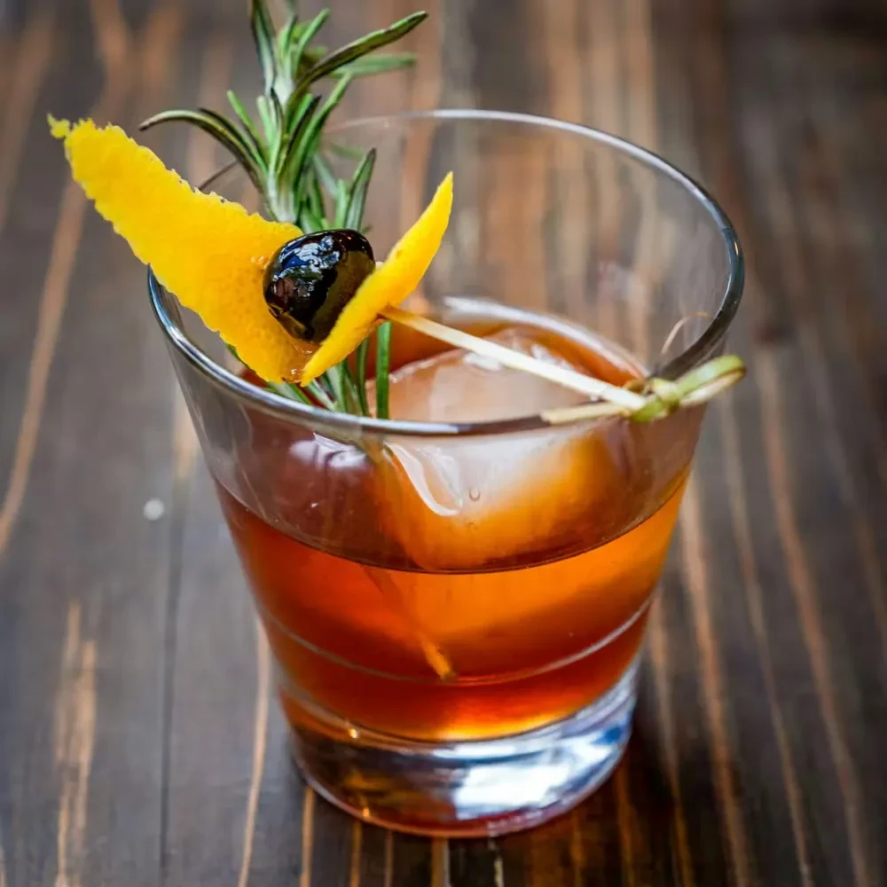 Amber cocktail with orange peel, cherry, rosemary, and ice cube.