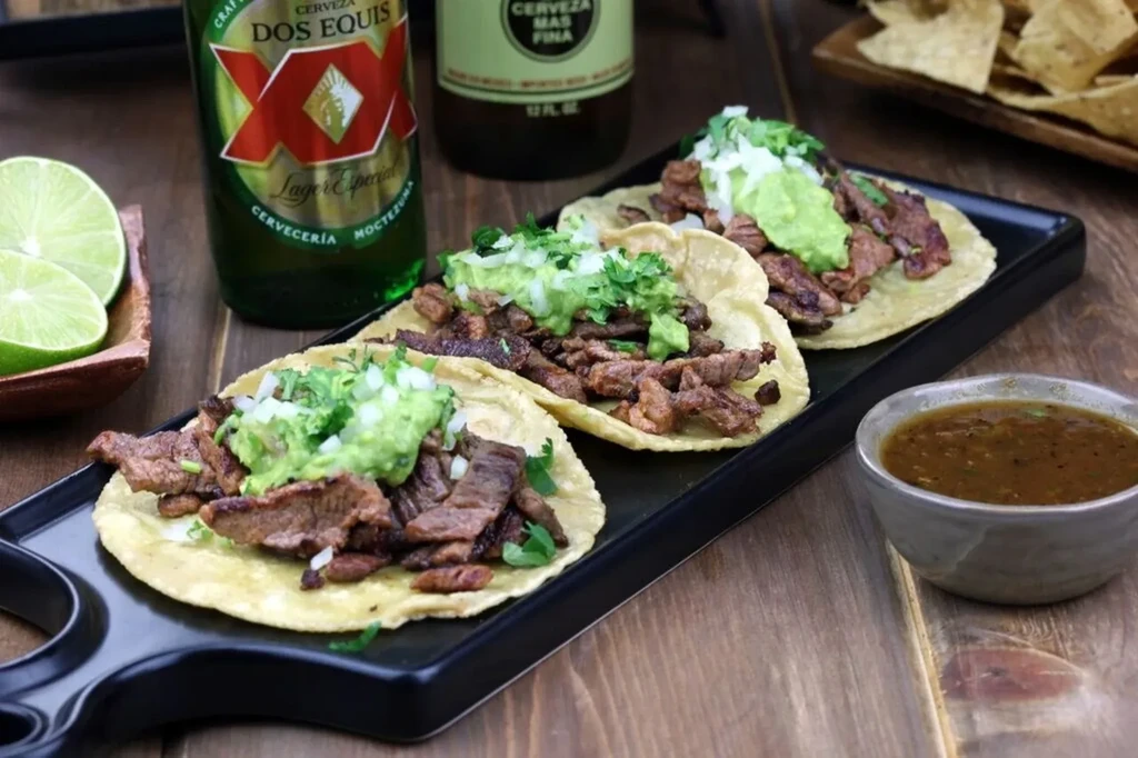 Close-up of three carne asada tacos with guacamole, lime, and salsa.
