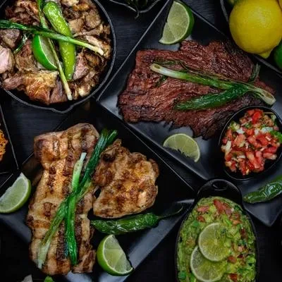 Overhead shot of grilled meats, guacamole, and salsa on dark dishes.