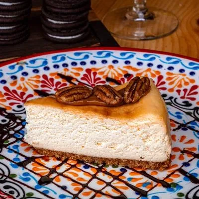 Cheesecake slice with caramel, pecans, and chocolate drizzle on patterned plate.