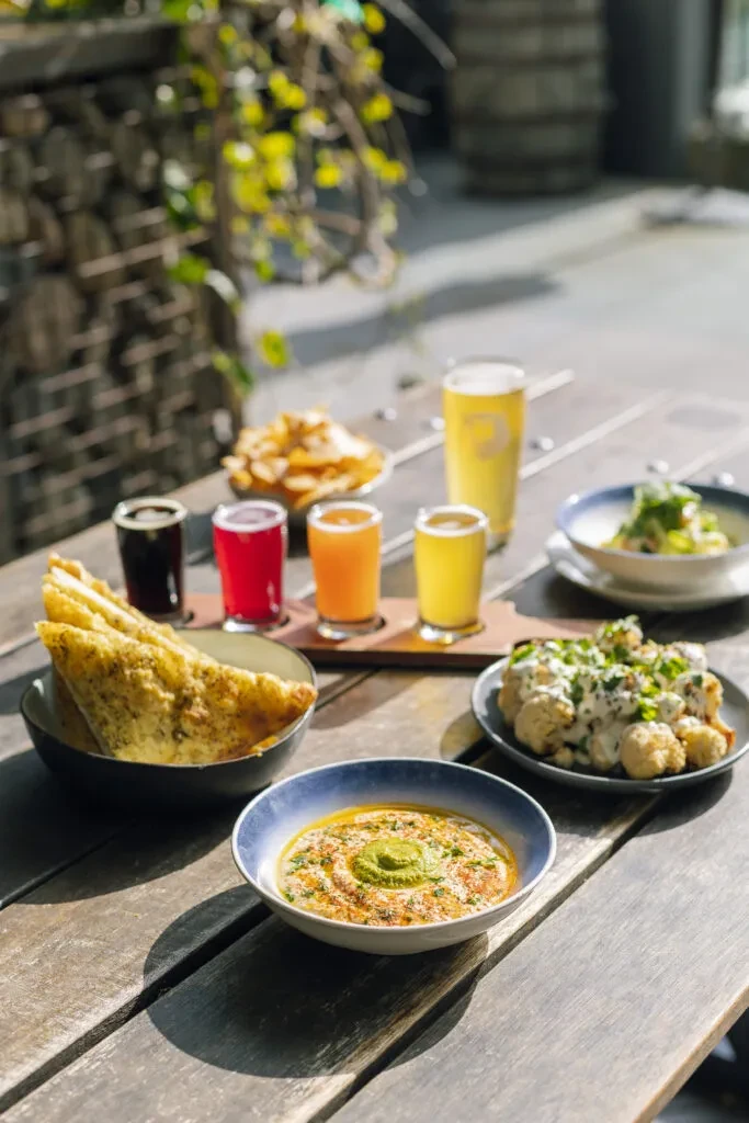 Sunny outdoor table with hummus, roasted cauliflower, flatbread, chips, and a beer flight.