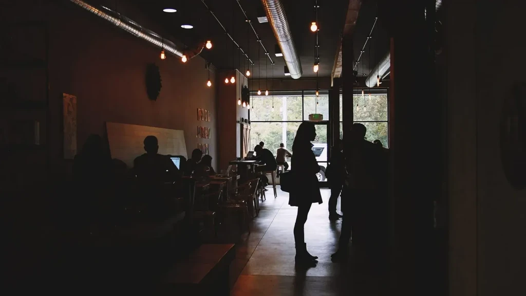 Low-lit industrial-chic cafe with strong silhouettes of people by windows.