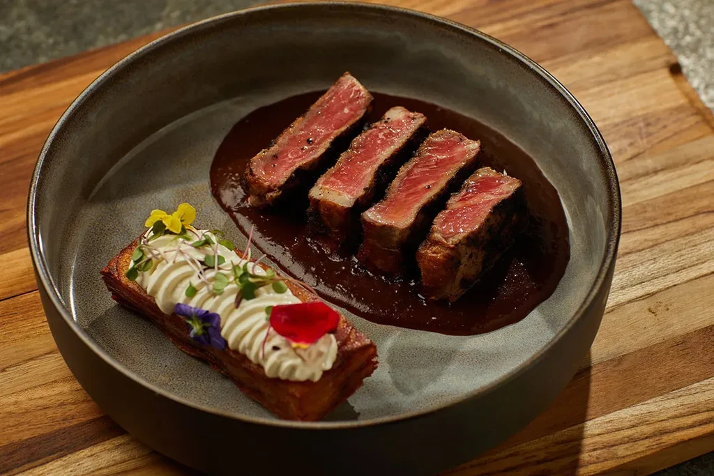 Plated medium-rare steak, creamy side, edible flowers, and microgreens.