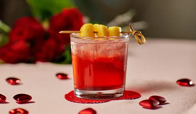 A red cocktail garnished with fruit, ice, red hearts, and roses.