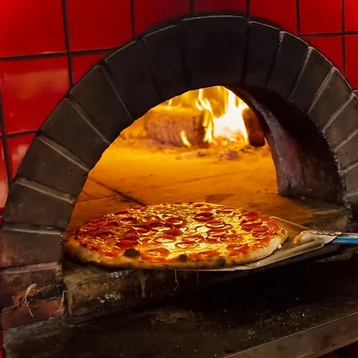 Cooked pepperoni pizza removed from a glowing brick wood-fired oven.