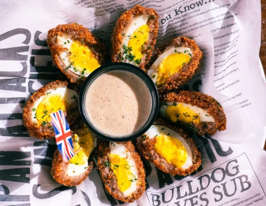Halved Scotch eggs, sauce, herbs, and Union Jack pick on newspaper liner.