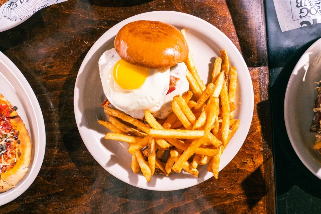 Burger with fried egg, fries, and pizza on a dark wooden table.