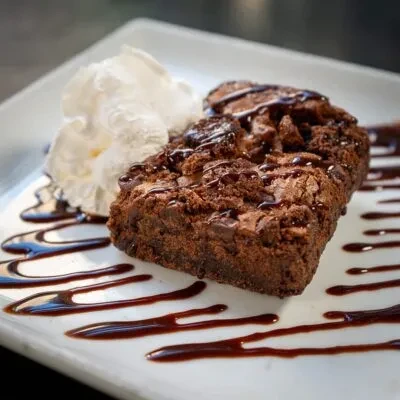 Rectangular brownie with chocolate sauce, whipped cream, decorated plate.