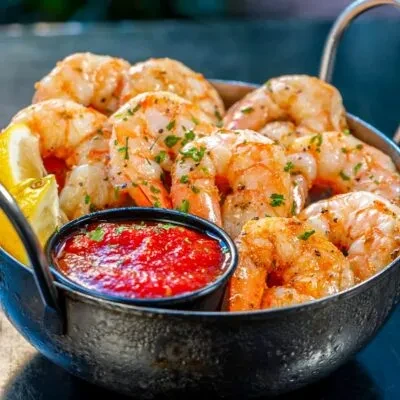 Seasoned cooked shrimp with red sauce and lemon in a dark bowl.
