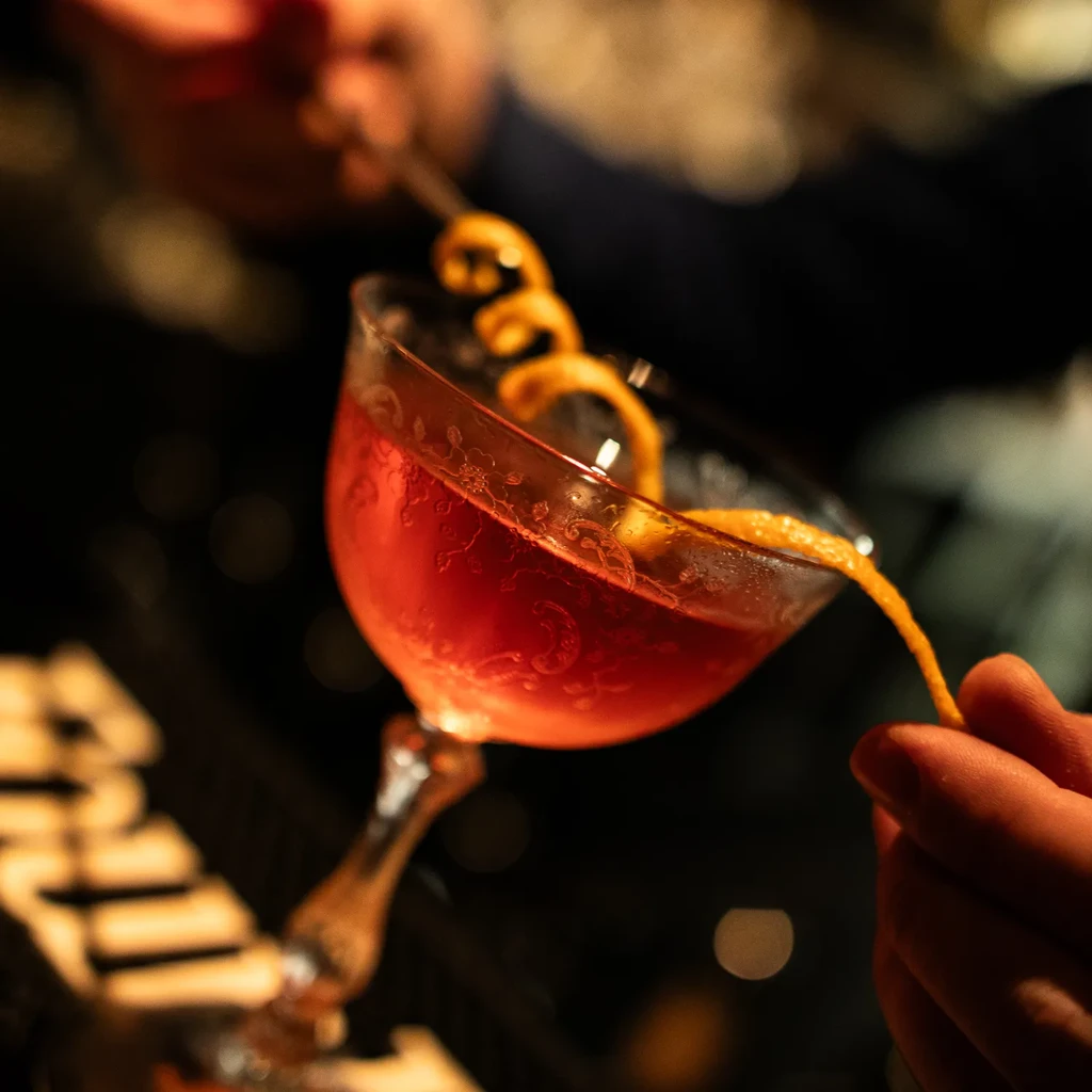 Hands garnish a red cocktail in an ornate glass with orange peel.