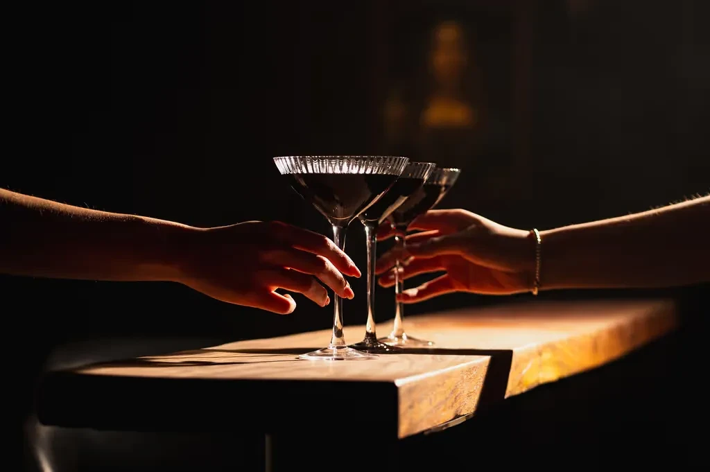 Three dark drinks on a wooden bar, hands reaching for a toast.