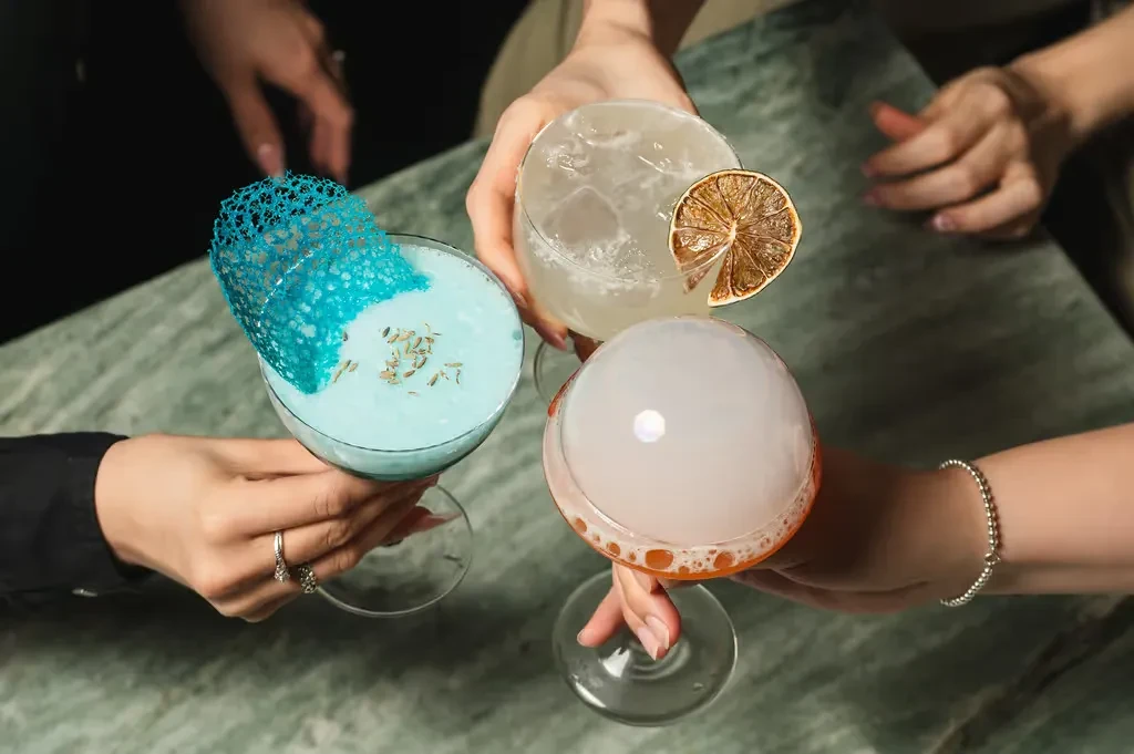 Three distinct, elaborately garnished cocktails held by hands on a table.