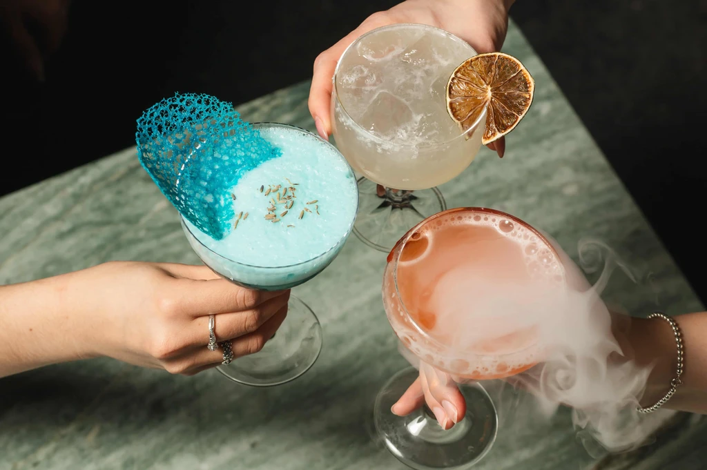Three hands hold three distinct, garnished cocktails, one smoking.