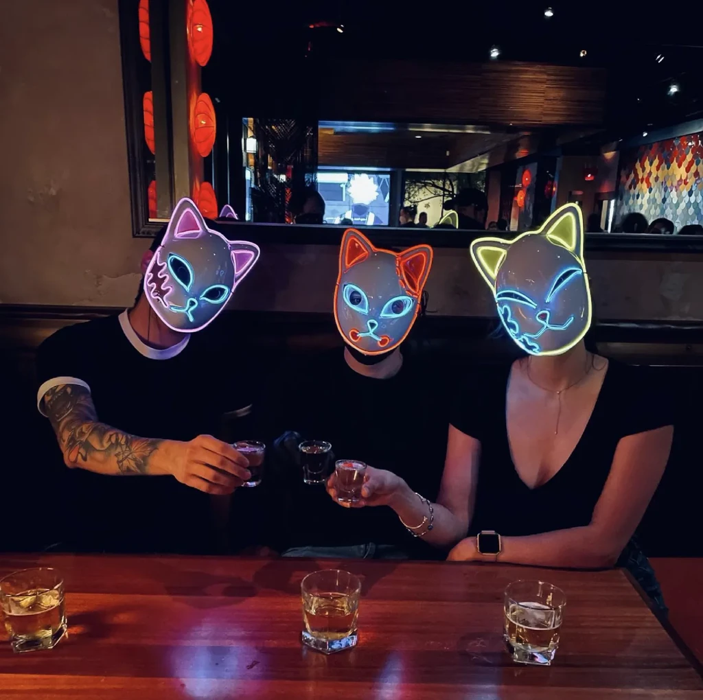 Three people in colorful glowing LED cat masks holding drinks.