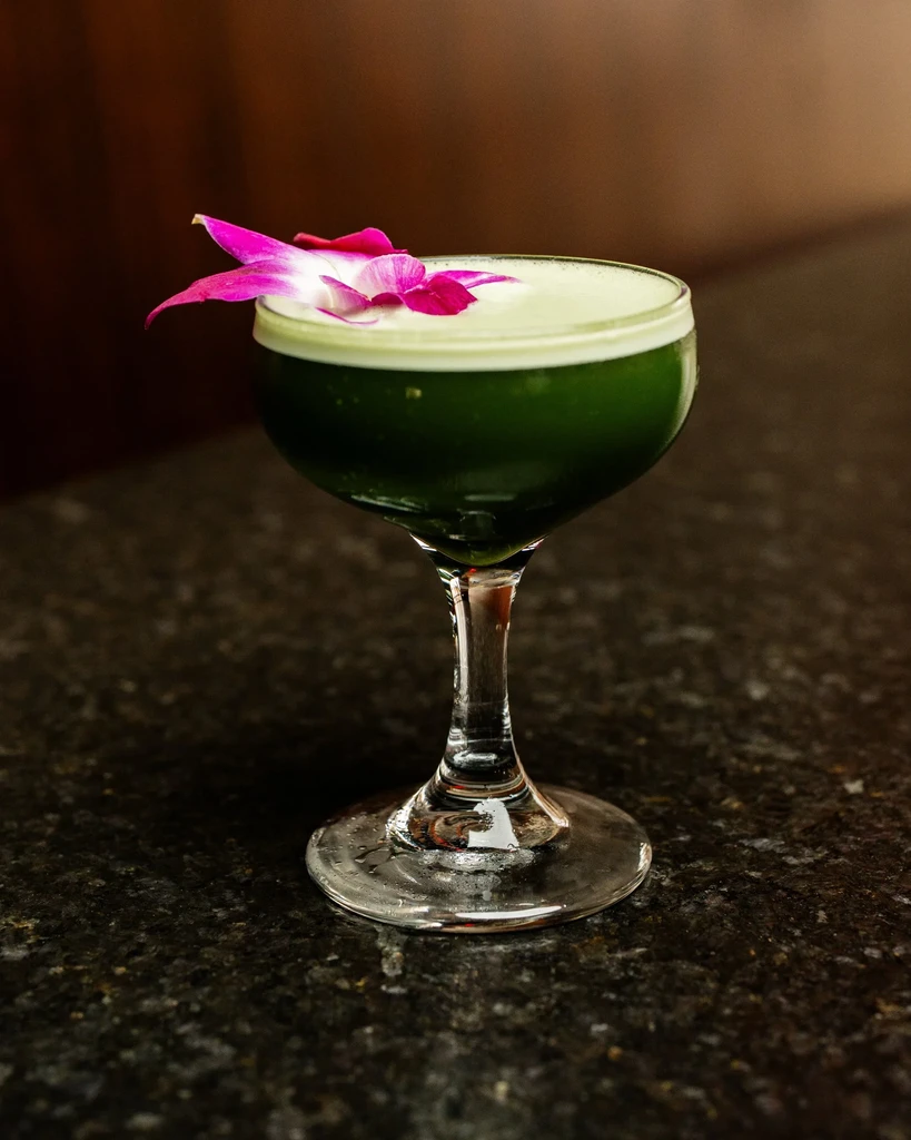 Green cocktail with white foam and a fuchsia orchid garnish.