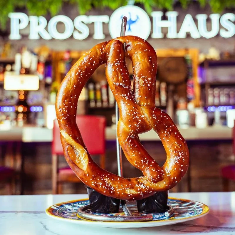 Large salted pretzel displayed before a "PROST HAUS" neon sign.