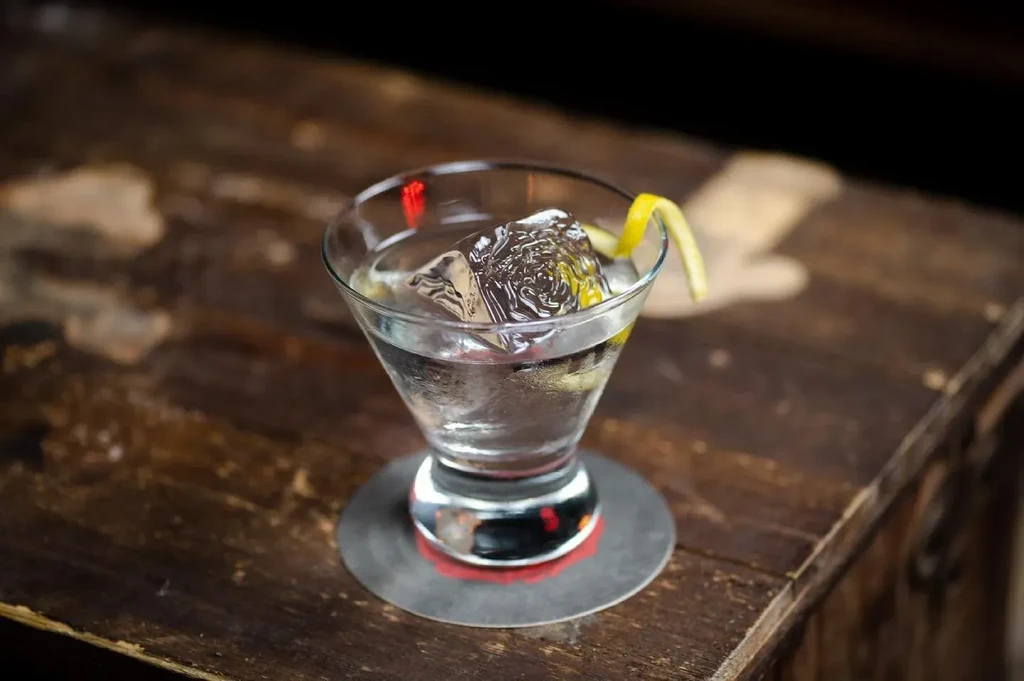 Clear cocktail with carved ice and lemon peel in a martini glass.