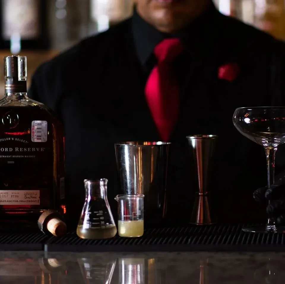Bourbon bottle, scientific glassware, and cocktail tools on a bar with blurred bartender.