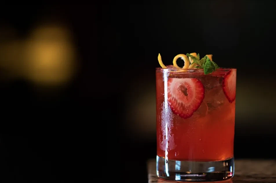 Red cocktail with strawberries, ice, orange peel, and mint garnish.