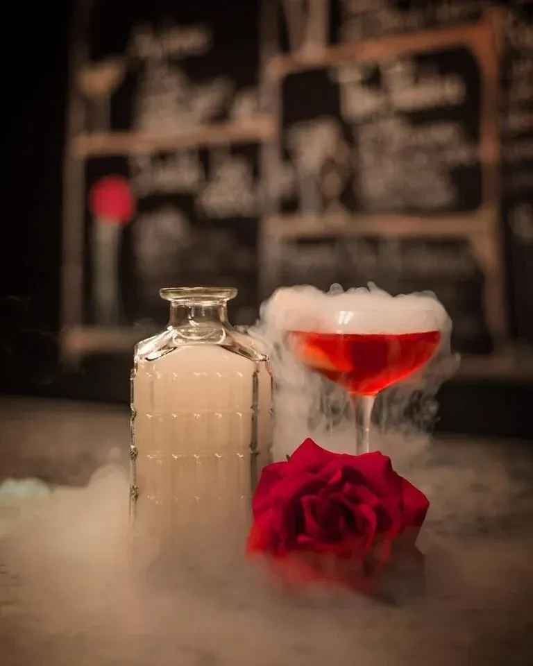 Low-light red cocktail, bottle, and rose in thick white vapor.