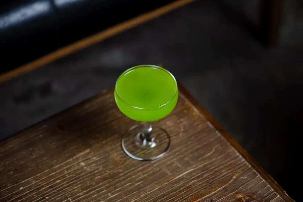 A vibrant green cocktail in a coupe glass on a dark wooden table.