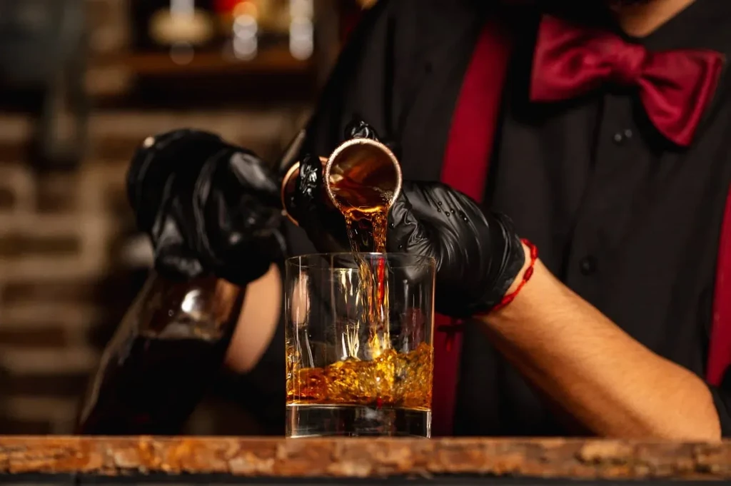 Bartender pours whiskey from copper jigger into glass with ice.
