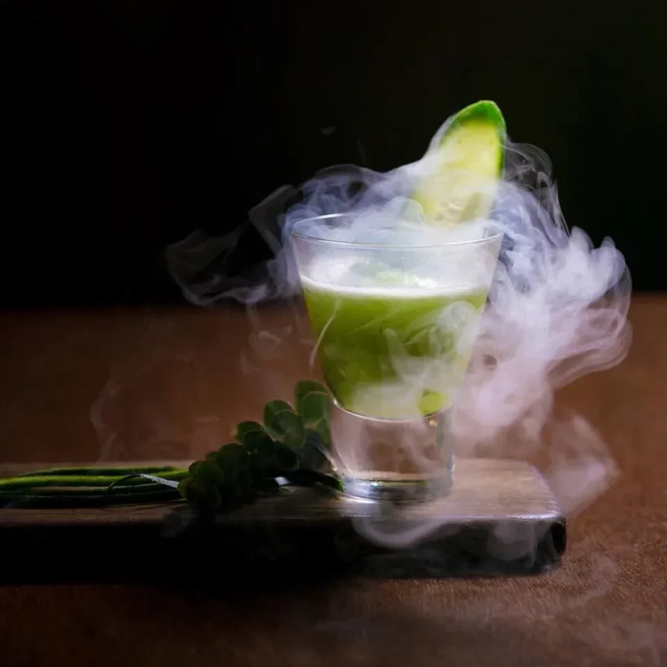 Green drink in clear glass, dry ice smoke, lime garnish.