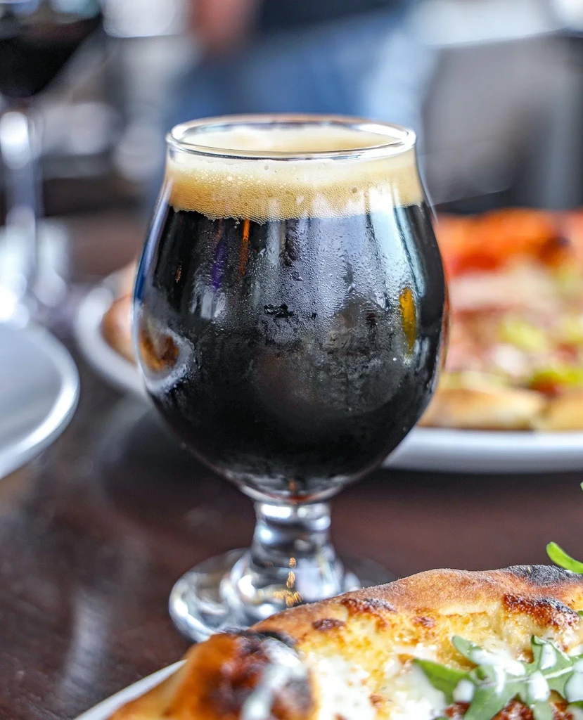 Dark beer in condensation-covered stemmed glass with pizza and creamy head.