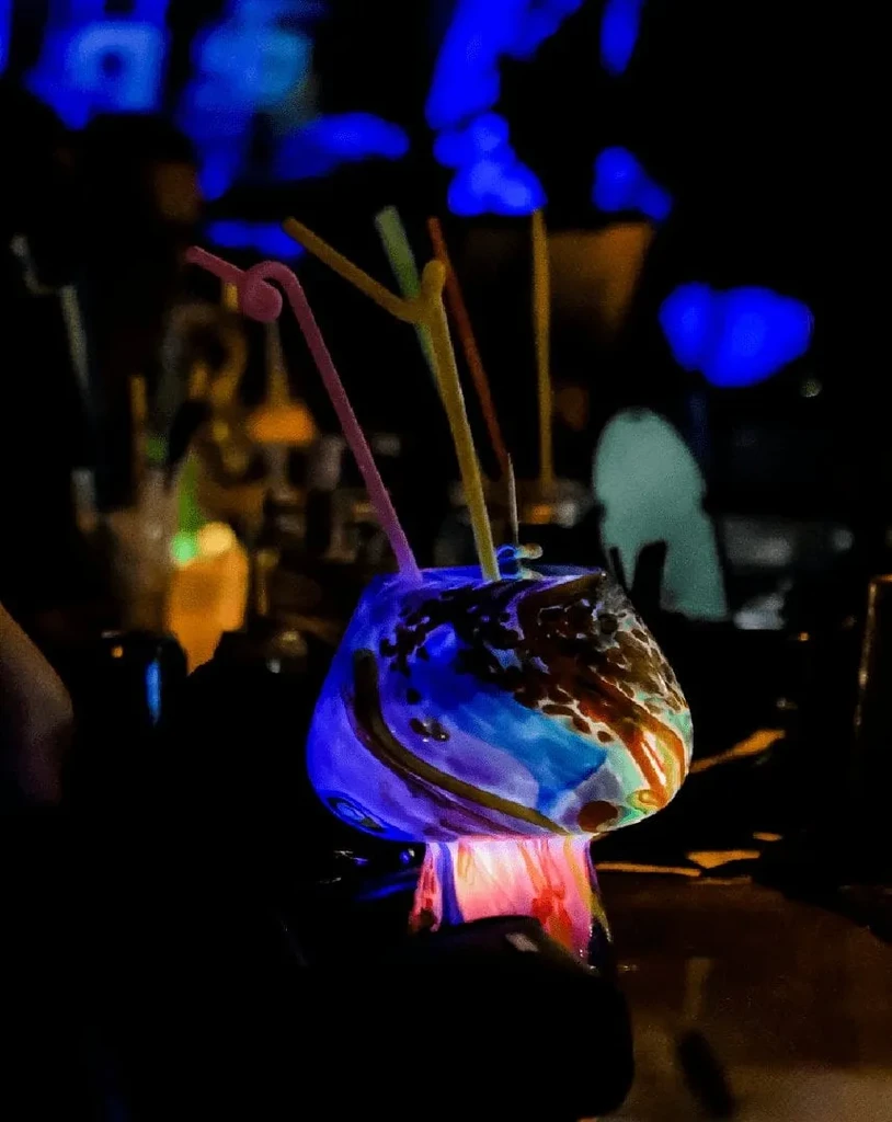 Colorful, illuminated drink in swirled glass with straws, dark background.