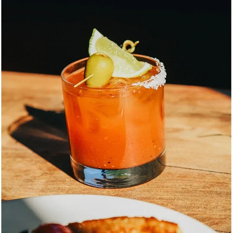 Reddish-orange cocktail with lime wedge, green olive, and salted rim.