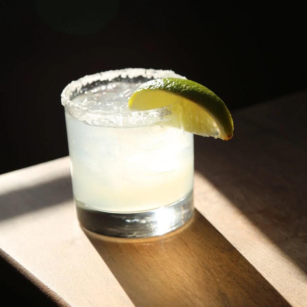 Brightly lit, salt-rimmed cocktail with lime and ice on dark background.