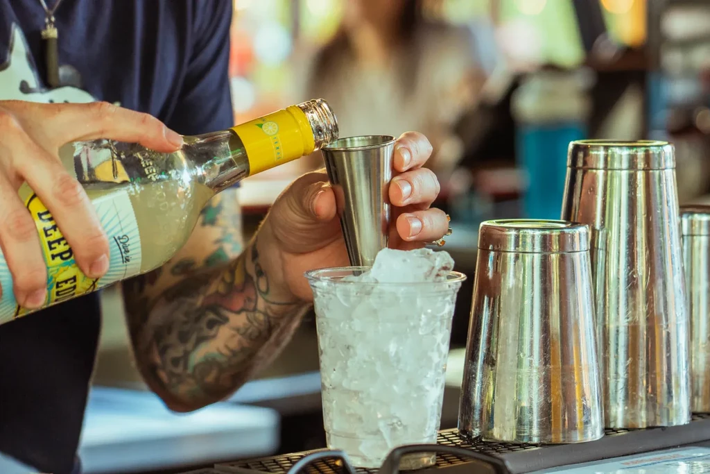 Tattooed bartender's hands pour from bottle into jigger and ice cup.