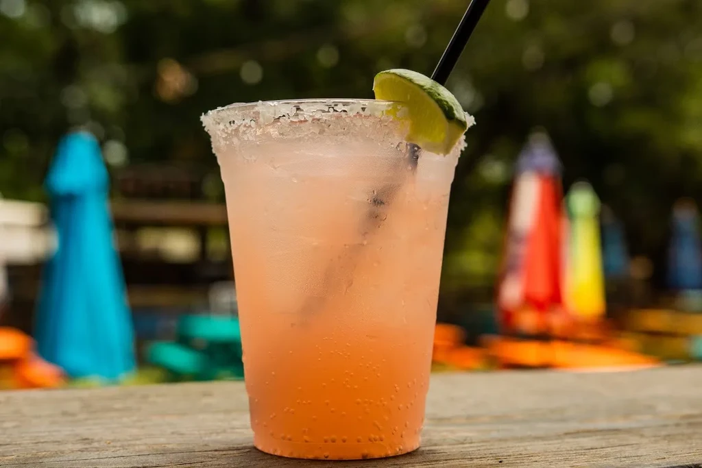 Pinkish-orange iced drink with salted rim, lime wedge on wood.