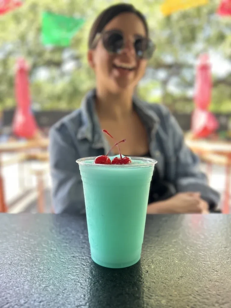 Blue-green frozen drink with cherries and condensation, person smiling outdoors.