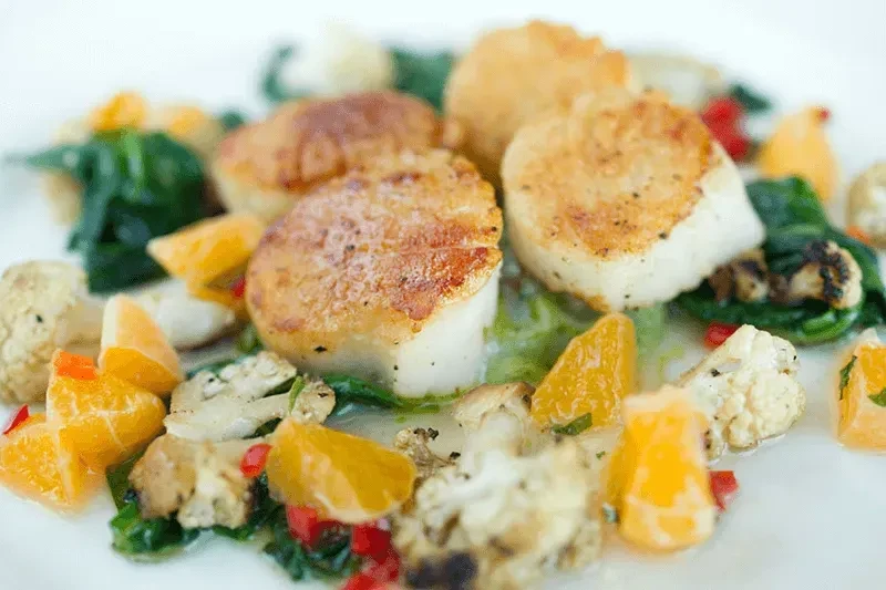 Plated seared scallops on spinach with orange, cauliflower, and red specks.