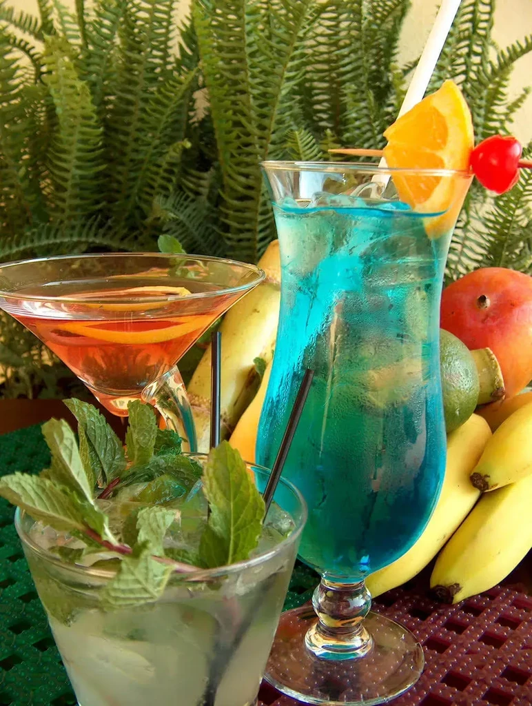 Three garnished tropical drinks and fresh fruits on a dark mat.