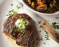 Seared steak with melting butter, green herbs, knife, and roasted Brussels sprouts.
