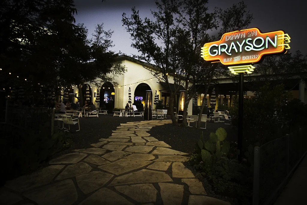 Night view of Down On Grayson restaurant with bright neon sign and patio.