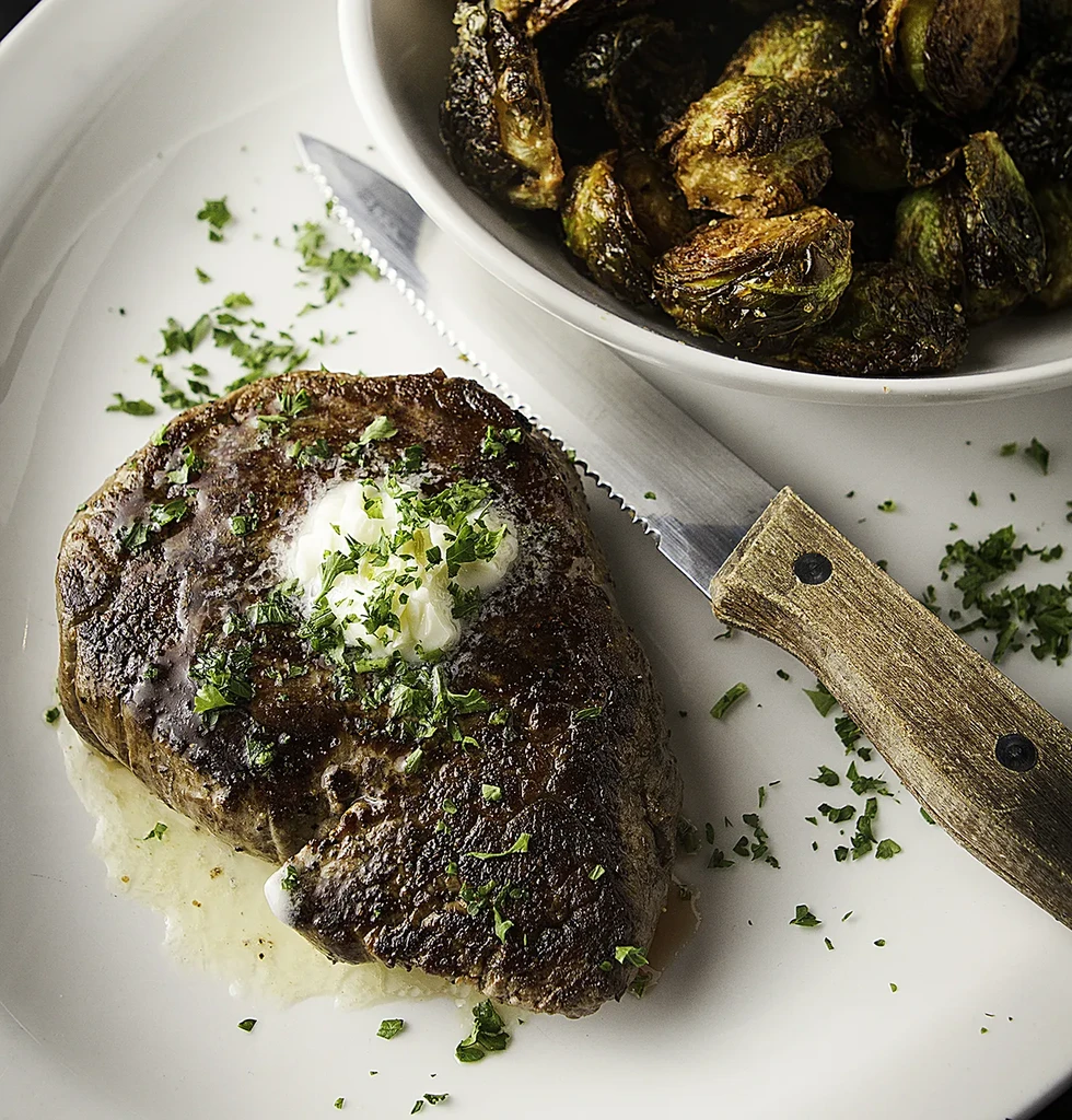 Close-up: Seared steak with melting butter, parsley, roasted brussels sprouts, knife.