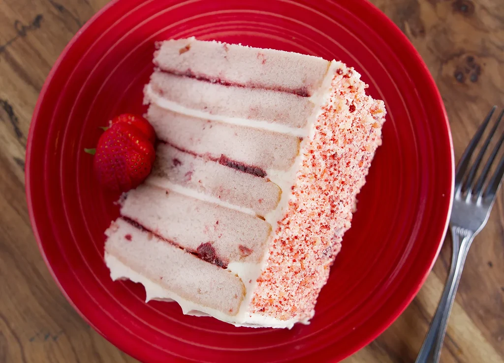 Strawberry layer cake slice, sprinkles, whole strawberry, fork, red plate.