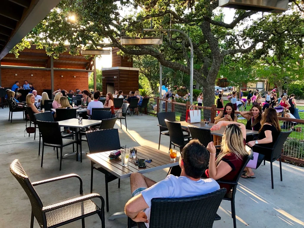 Lively restaurant patio under oak trees, people socializing with drinks.